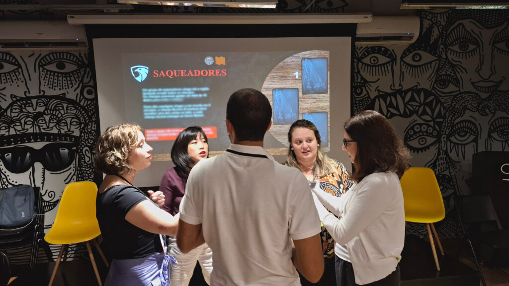 Grupo de participantes reunido em dinâmica presencial do jogo Falkenauge Sobrevivência, discutindo estratégias e tomando decisões colaborativas sob pressão.